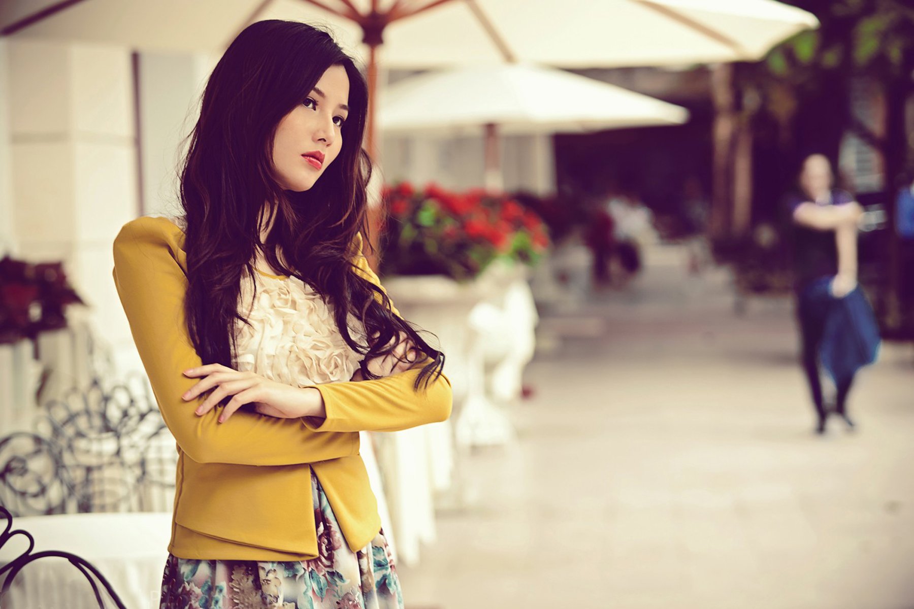 HD desktop wallpaper featuring an Asian woman with long hair, standing outdoors with arms crossed, looking thoughtful. Flowers and white umbrellas are in the background.