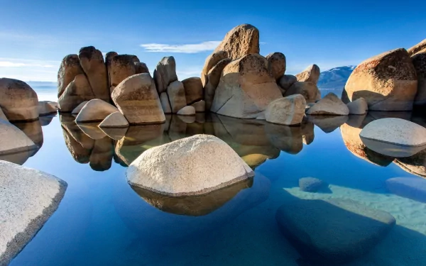 A serene lake reflects large, smooth rocks under a clear blue sky, creating a stunning nature scene captured in high-definition photography.