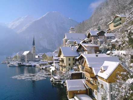 HD desktop wallpaper of a snow-covered Hallstatt town in winter, showcasing picturesque man-made buildings nestled alongside a serene lake, with a backdrop of snow-capped mountains.