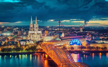 Nighttime HD desktop wallpaper of Cologne showcasing the illuminated Hohenzollern Bridge, Cologne Cathedral, and city skyline under a dramatic sky.