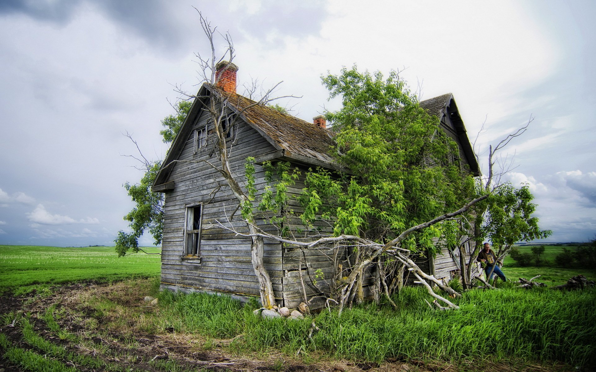 HD desktop wallpaper featuring an old, weathered man-made wooden cabin surrounded by green grass and trees under a cloudy sky.