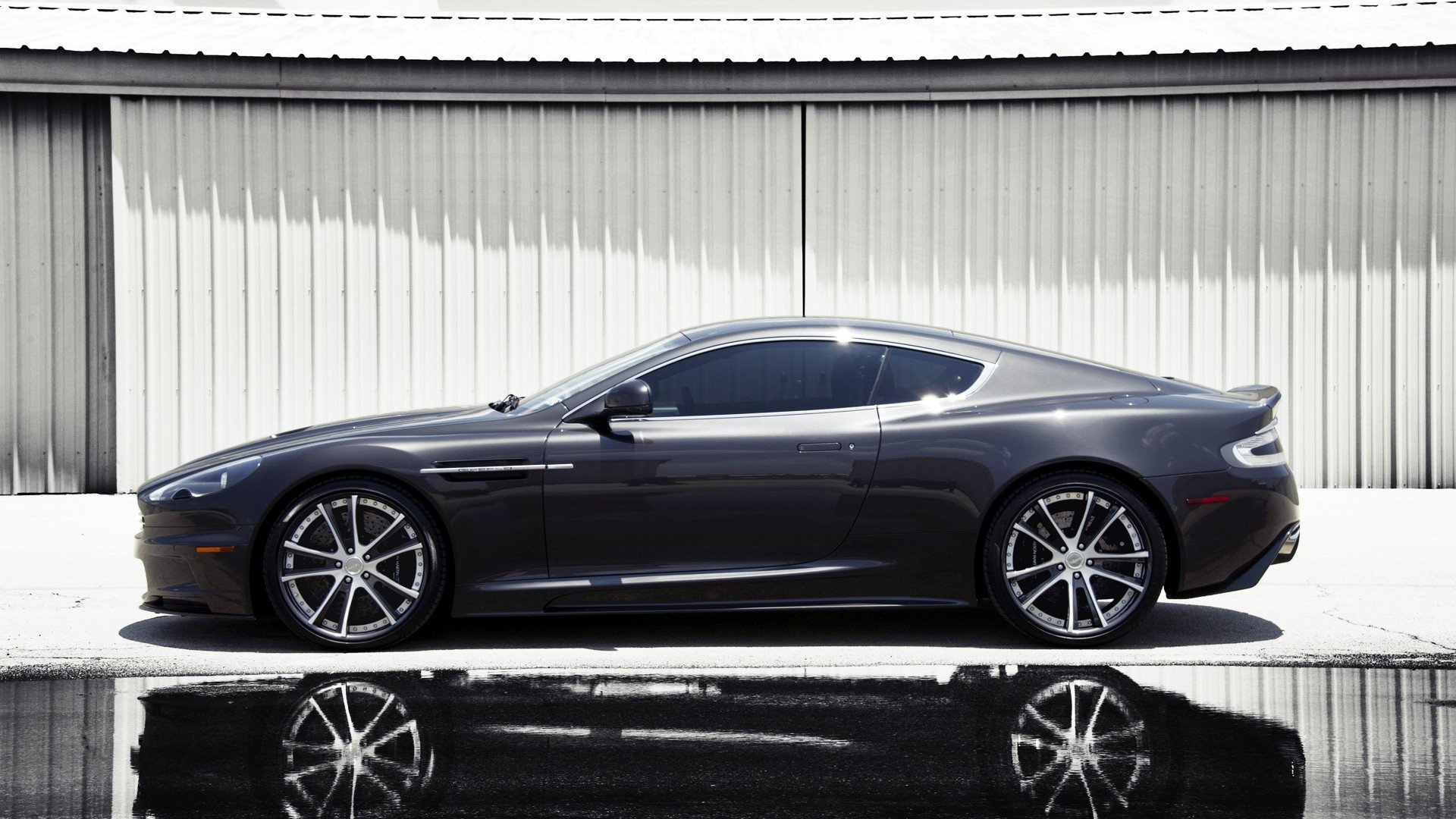 HD PC desktop wallpaper background: Aston Martin One-77 vehicle in dark side profile, parked before a corrugated white wall with a crisp reflection on wet pavement.