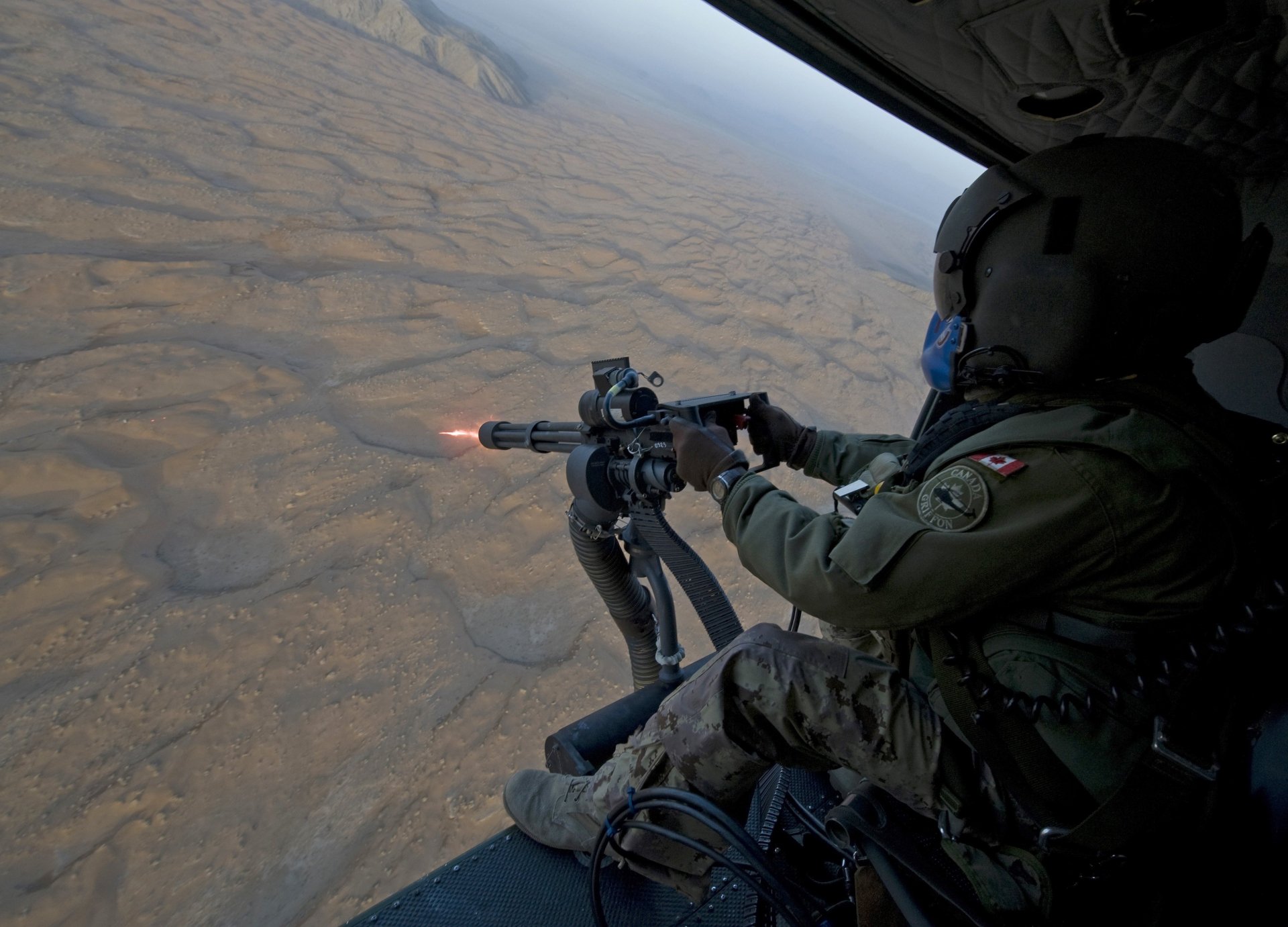 A high-definition wallpaper featuring a man in military gear operating a machine gun from a helicopter, showcasing action and intensity against a textured ground backdrop.