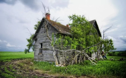 HD desktop wallpaper featuring an old, weathered man-made wooden cabin surrounded by green grass and trees under a cloudy sky.