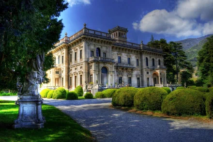 A stunning mansion surrounded by lush greenery and manicured hedges, set against a backdrop of blue skies and clouds. This high-definition image serves as an impressive desktop wallpaper.