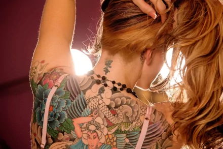 HD wallpaper featuring a woman with intricate, colorful tattoos on her back and neck, her hair pulled up in a ponytail, and light shining in the background.