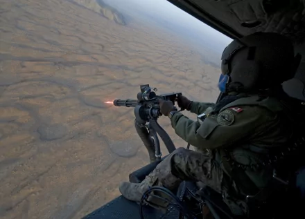 A high-definition wallpaper featuring a man in military gear operating a machine gun from a helicopter, showcasing action and intensity against a textured ground backdrop.