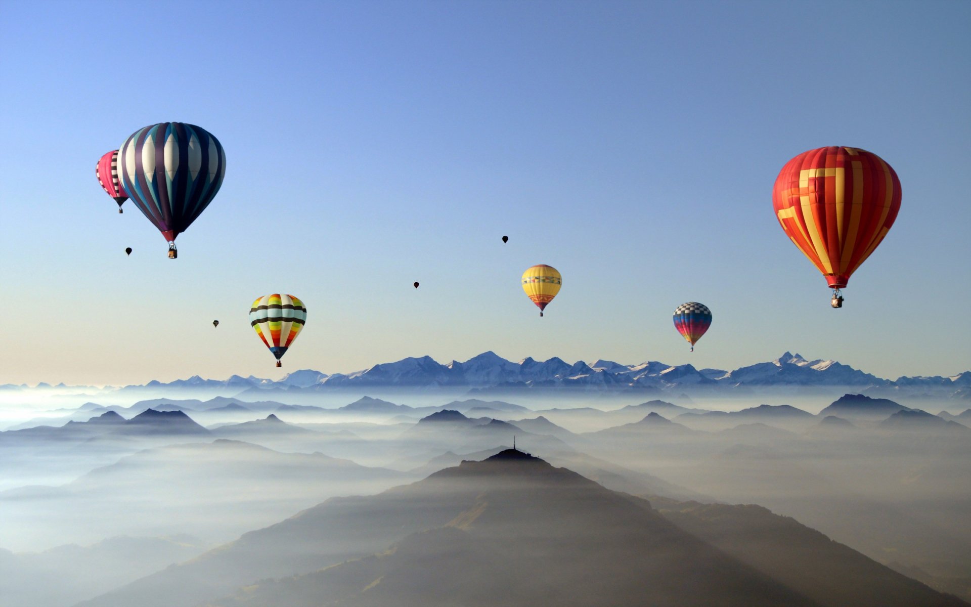 HD desktop wallpaper featuring colorful hot air balloons floating above misty mountain peaks under a clear blue sky.