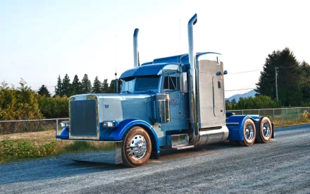 HD desktop wallpaper featuring a classic blue Peterbilt semi-truck parked on a rural road with trees and mountains in the background.