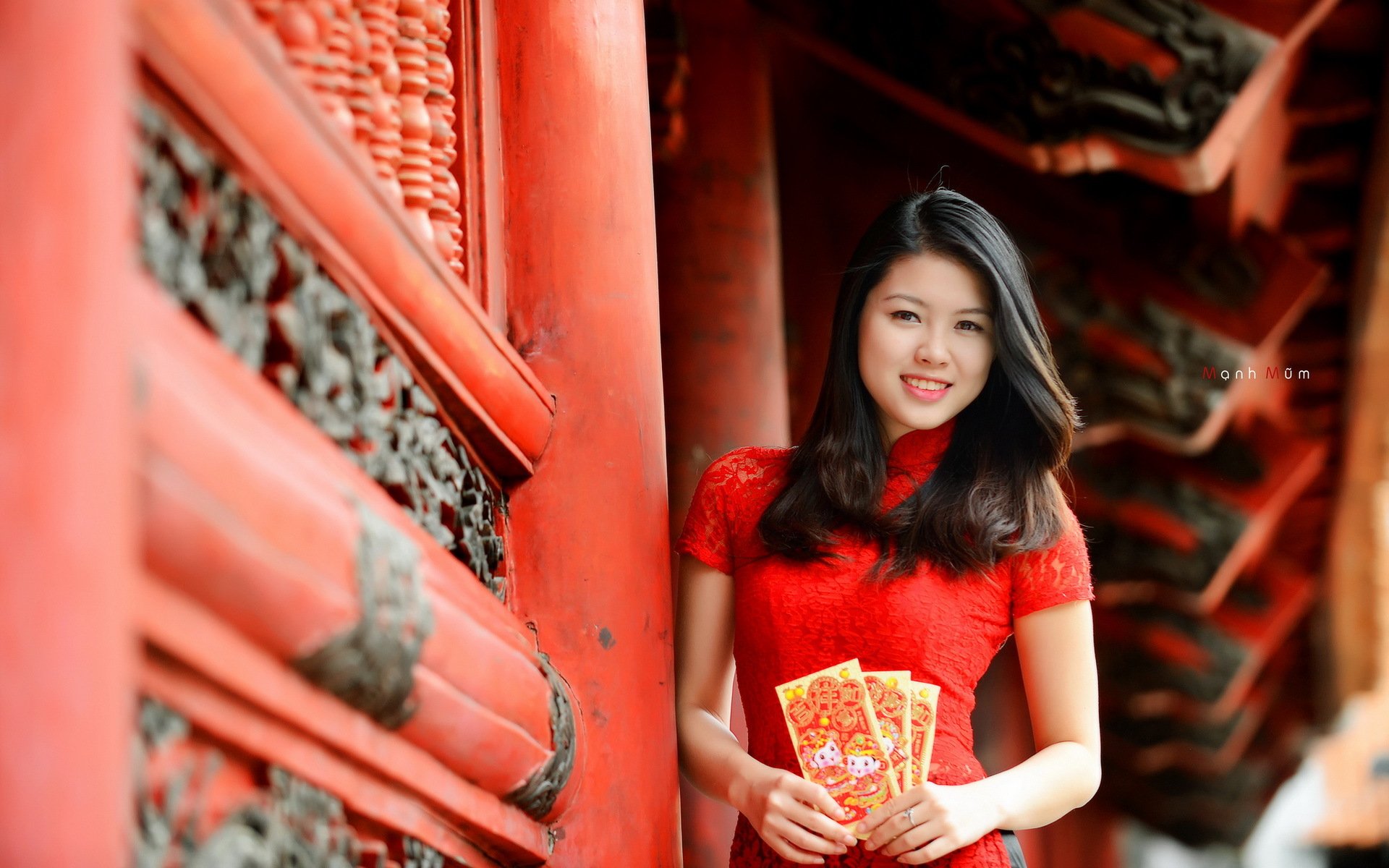 A smiling Asian woman in a red traditional outfit stands by a vibrant red wall, holding decorative envelopes, showcasing a festive atmosphere in this HD desktop wallpaper.