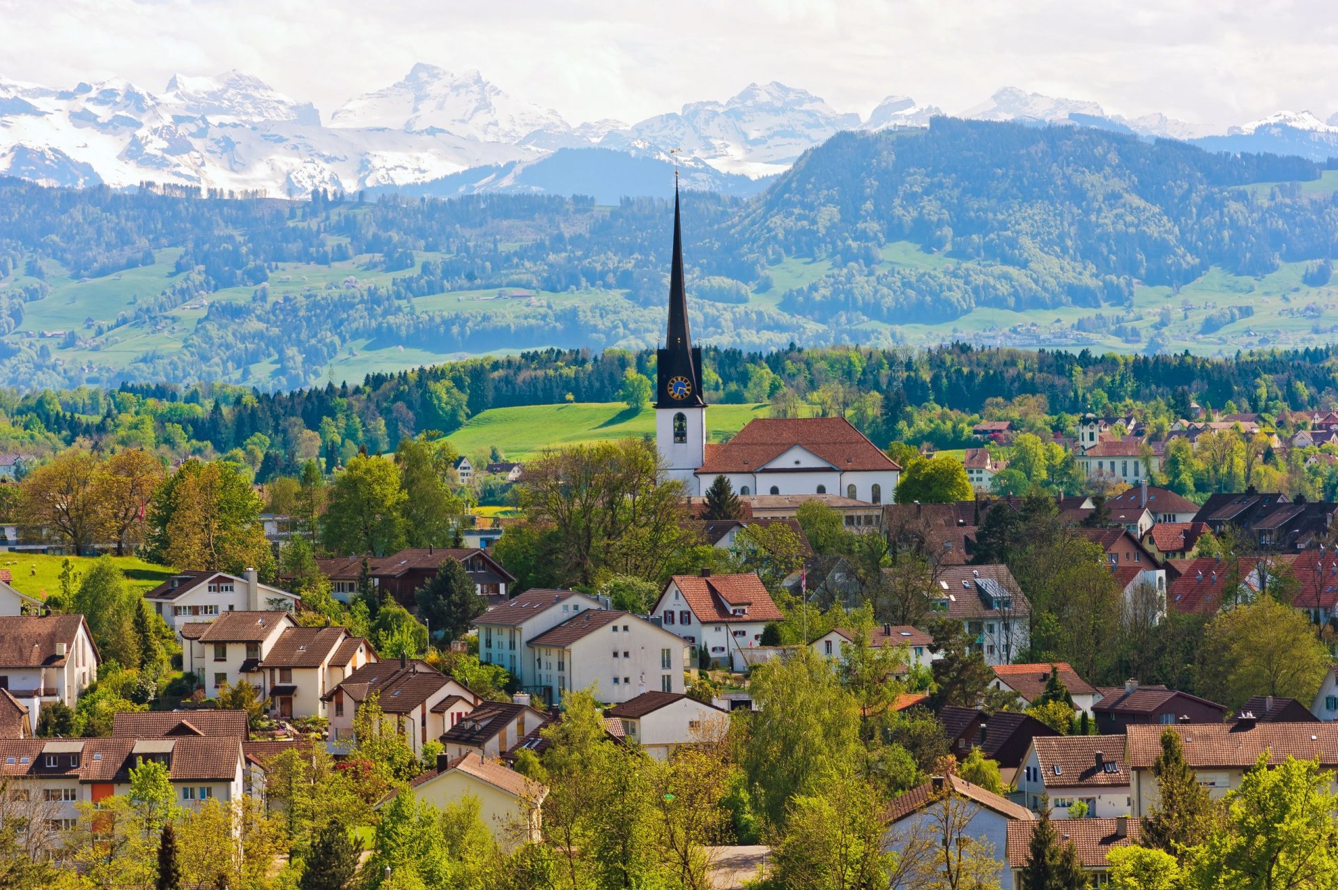 Panoramic HD desktop wallpaper featuring a Swiss village with charming houses, a prominent church, and mountainous backdrop under a clear sky.