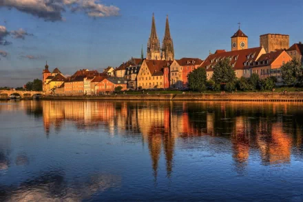 HD PC desktop wallpaper/background: man-made riverside village with red-roofed houses and twin church spires glowing in warm evening light, reflected on calm water.