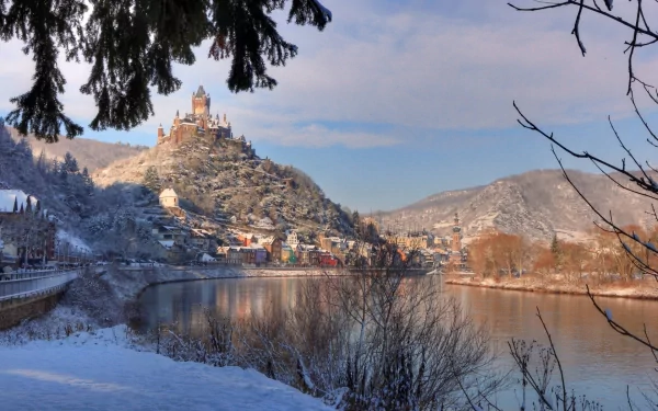 HD desktop wallpaper featuring Cochem Castle perched on a hill overlooking a river, framed by winter trees under a clear blue sky.