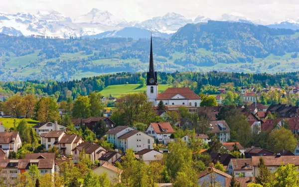 Panoramic HD desktop wallpaper featuring a Swiss village with charming houses, a prominent church, and mountainous backdrop under a clear sky.