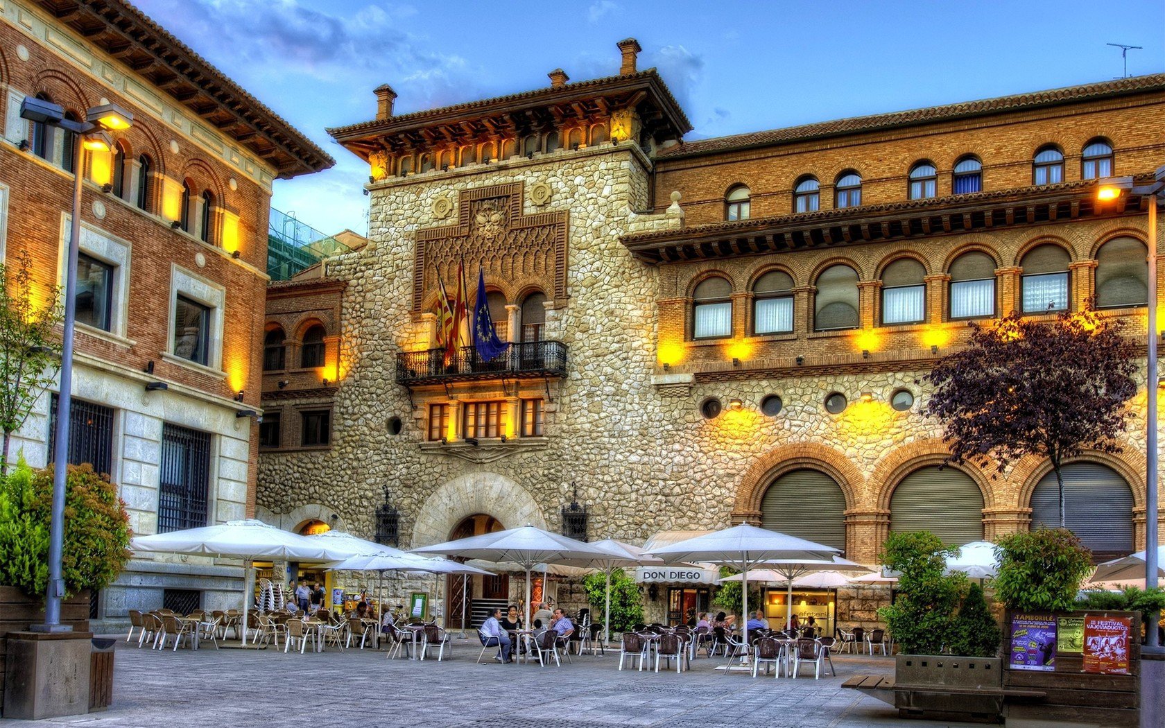HD desktop wallpaper showcasing a vibrant evening scene of man-made architecture in Teruel, with illuminated historic buildings and outdoor seating under a clear sky.