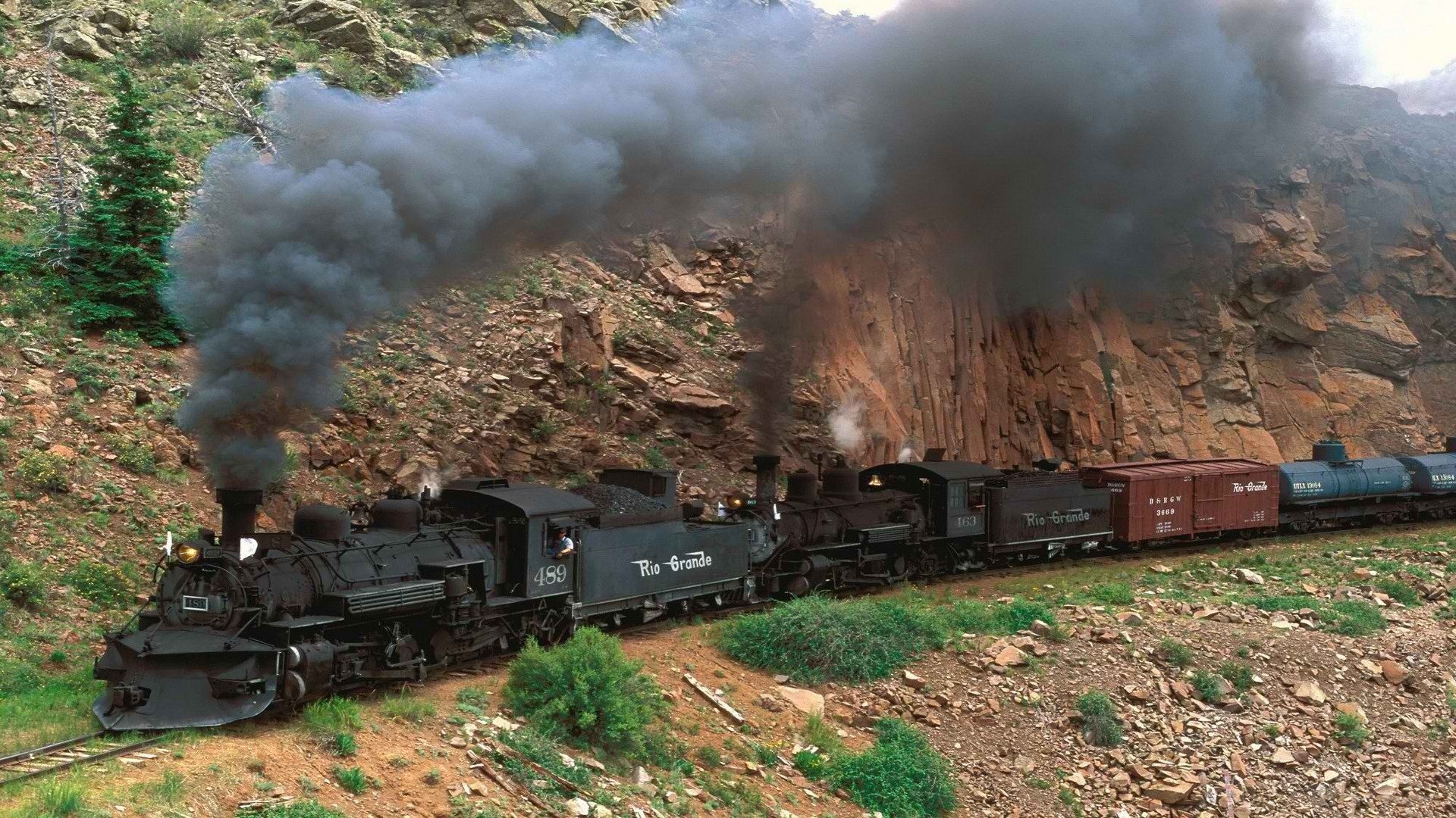 HD desktop wallpaper featuring a powerful locomotive emitting thick black smoke as it moves through a rugged, rocky landscape surrounded by greenery.