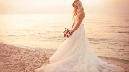 HD desktop wallpaper of a bride in a flowing white gown holding a bouquet, standing on a beach at sunset with calm water in the background.