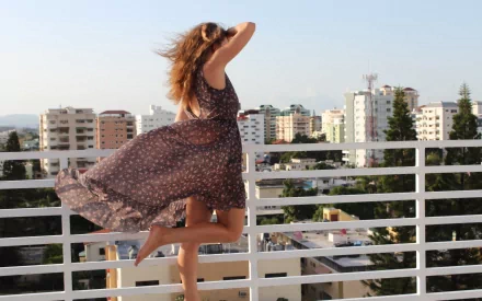A woman with long hair, seen from the rear, stands on a balcony overlooking a cityscape. She wears a flowing dress that catches the wind. HD desktop wallpaper and background.
