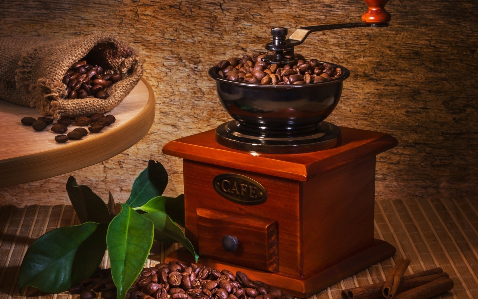 HD desktop wallpaper of a vintage coffee grinder filled with coffee beans, surrounded by spilled beans and green leaves on a rustic wooden surface.