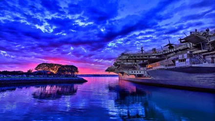 A stunning HD wallpaper featuring a military aircraft carrier reflected in calm waters, set against a vibrant sunset with dramatic clouds.