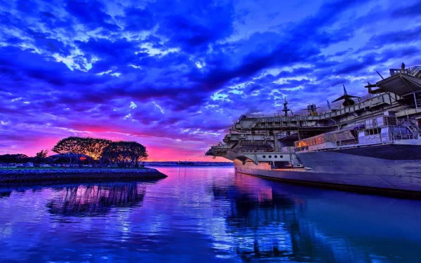 A stunning HD wallpaper featuring a military aircraft carrier reflected in calm waters, set against a vibrant sunset with dramatic clouds.