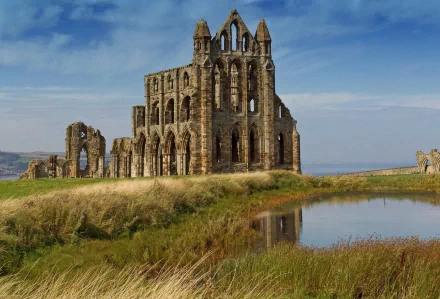 HD PC desktop wallpaper featuring a striking man-made ruin set against a blue sky, reflected in a calm pond surrounded by lush grass.