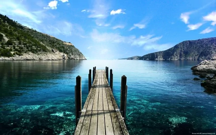 HD desktop wallpaper featuring a man-made wooden pier extending into clear, calm blue waters surrounded by rocky cliffs under a bright sky.