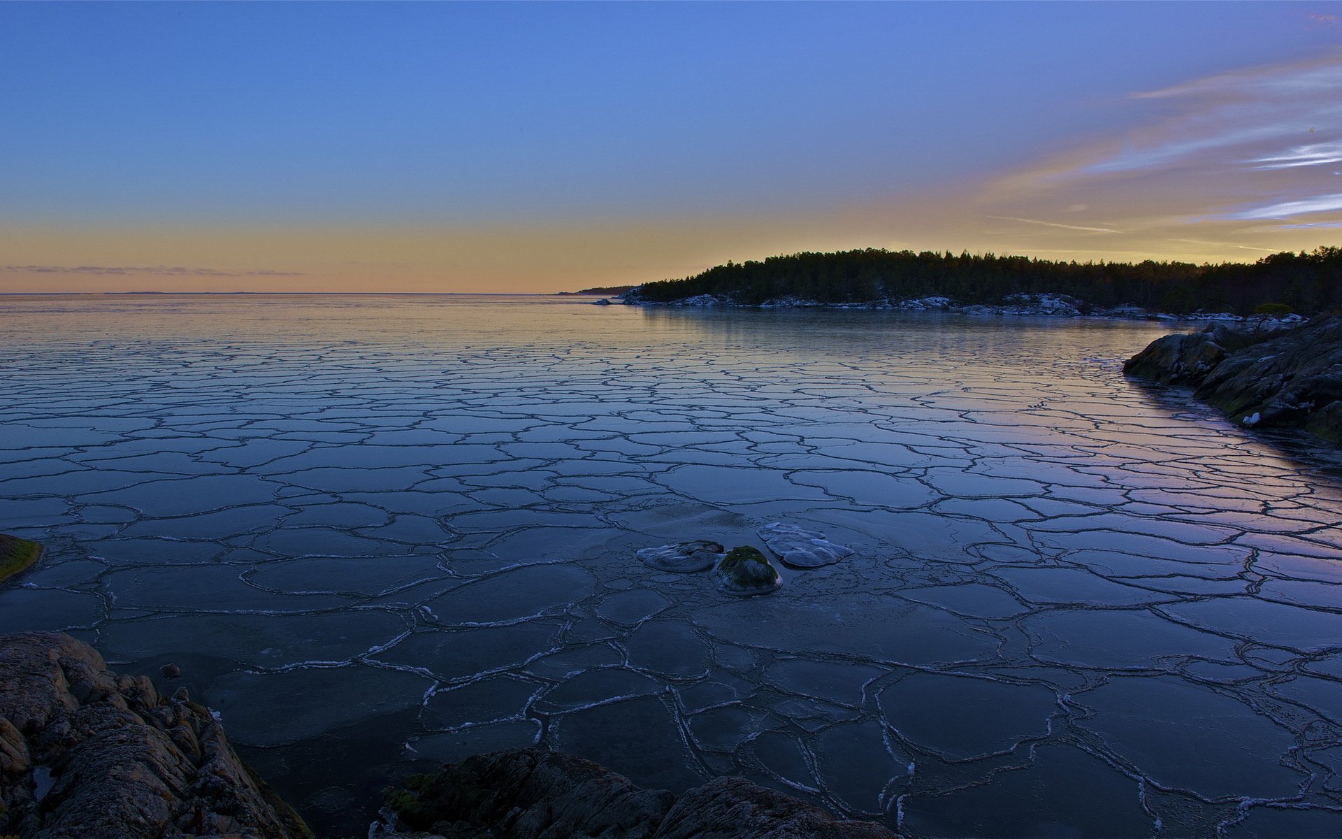 HD PC desktop wallpaper of a nature scene: cracked sea ice stretching over calm coastal water at dusk, with a silhouetted tree-lined shoreline under a pastel sky.