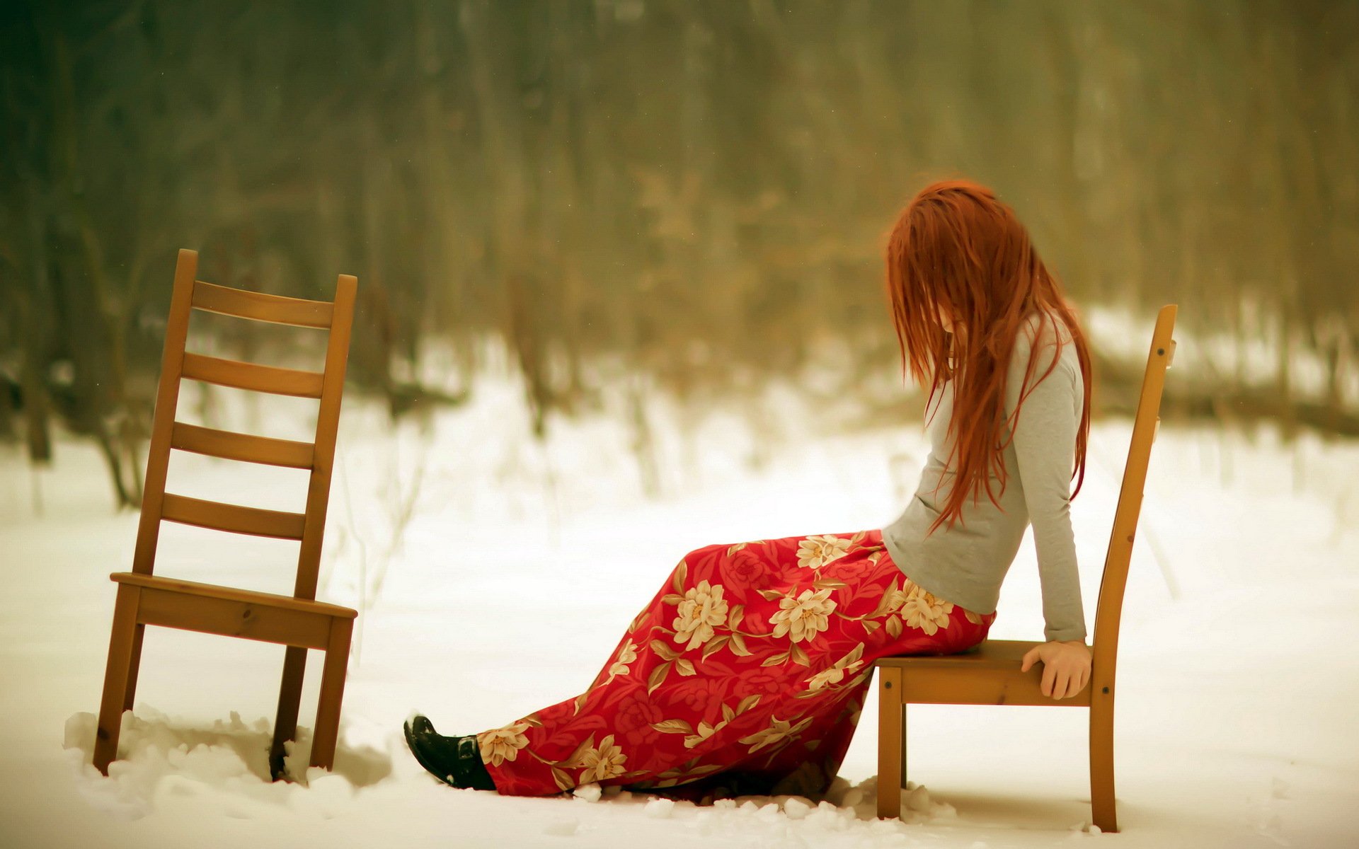HD PC desktop wallpaper of a woman with long red hair sitting on a chair in the snow, wearing a floral skirt, conveying a contemplative mood.