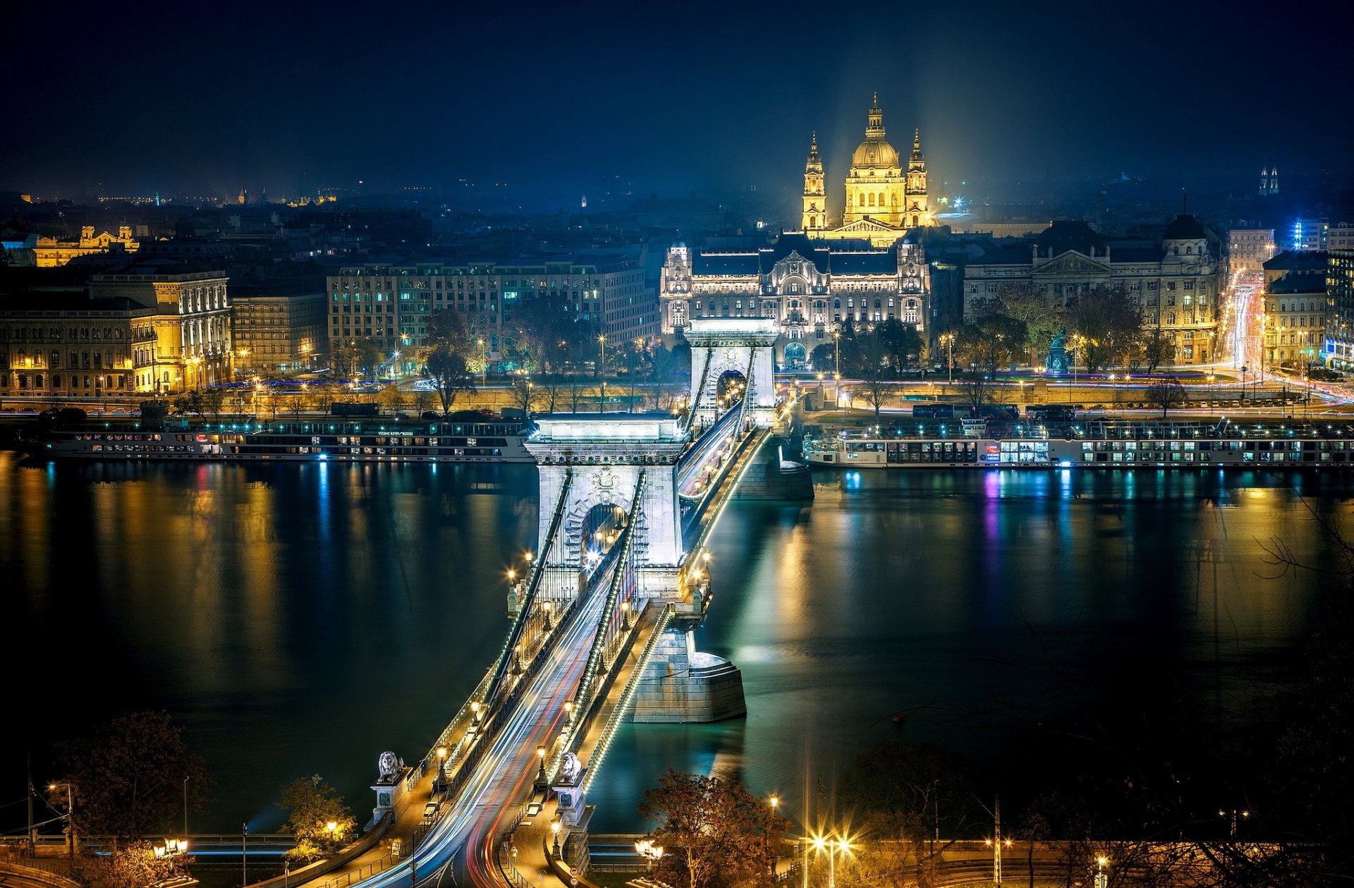 A stunning night view of Budapest's Chain Bridge, illuminated against the backdrop of the cityscape, featuring reflections on the river and the grandeur of nearby buildings.