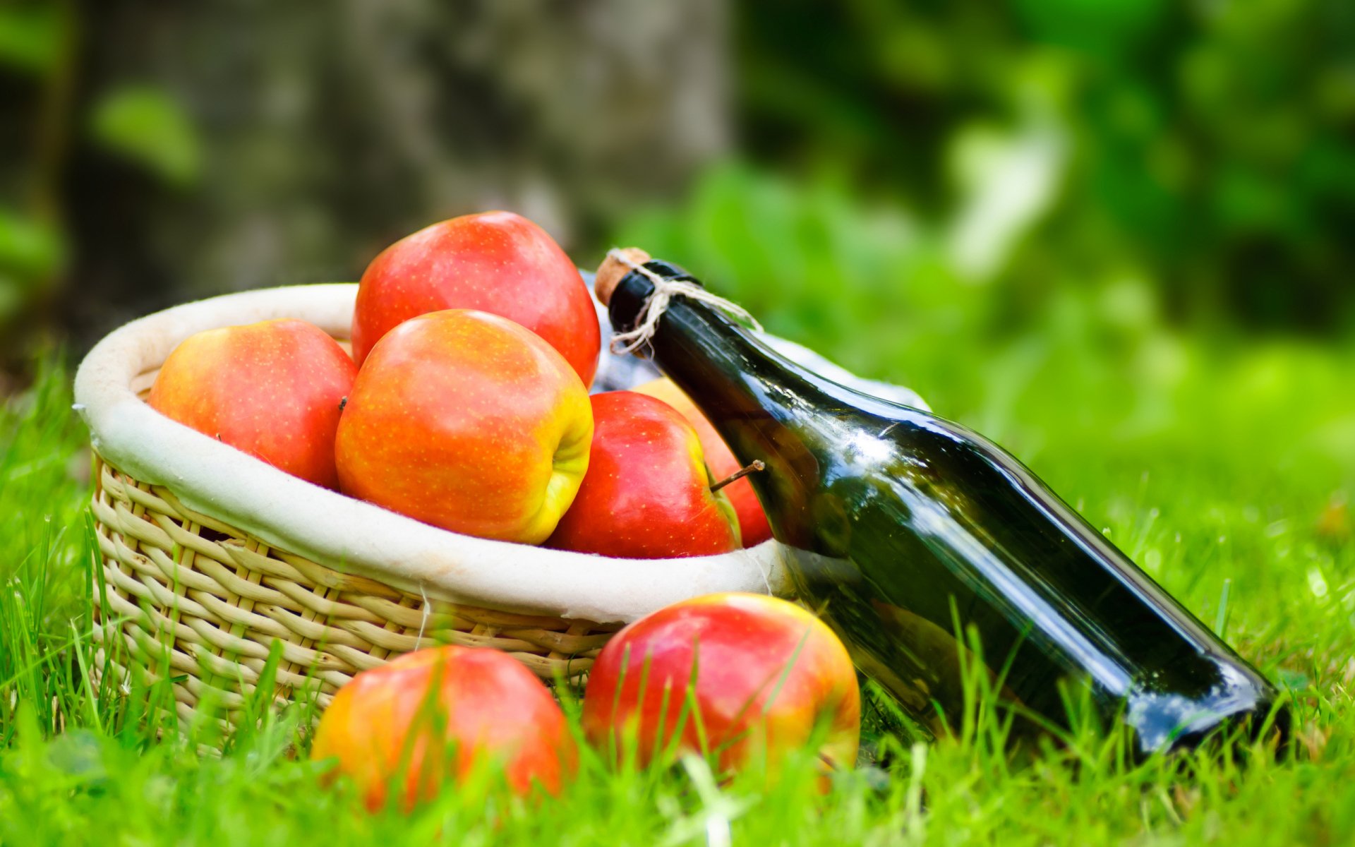 2K Quad HD PC desktop wallpaper: basket of red apples and a dark glass bottle on grass with a blurred green background — food, apple.