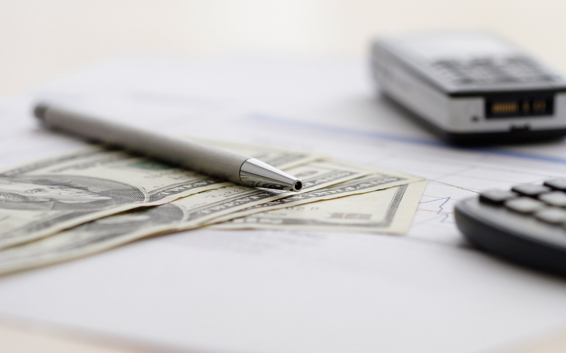 Macro photography of a pen resting on US dollar bills with a calculator and a phone blurred in the background, captured as an HD PC desktop wallpaper and background.