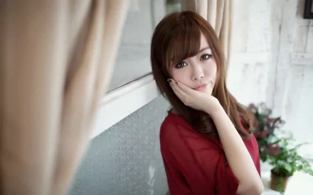 Asian woman in a red top, touching her face, stands in front of a window with beige curtains in an HD desktop wallpaper and background. The setting includes potted plants and light-colored walls.