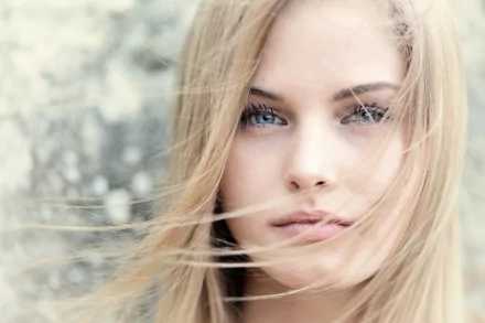 HD desktop wallpaper featuring a close-up of a woman with blonde hair and blue eyes, with loose strands of hair gently blowing across her face, against a softly blurred background.