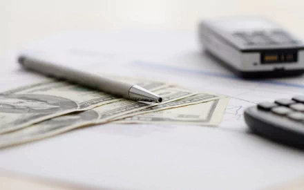 Macro photography of a pen resting on US dollar bills with a calculator and a phone blurred in the background, captured as an HD PC desktop wallpaper and background.