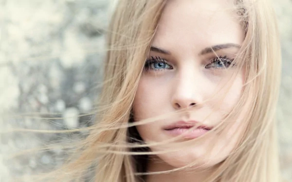 HD desktop wallpaper featuring a close-up of a woman with blonde hair and blue eyes, with loose strands of hair gently blowing across her face, against a softly blurred background.
