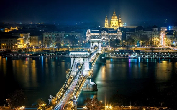 A stunning night view of Budapest's Chain Bridge, illuminated against the backdrop of the cityscape, featuring reflections on the river and the grandeur of nearby buildings.