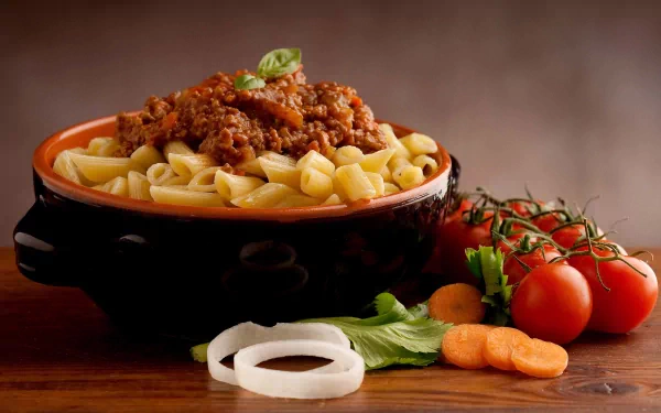 A delicious bowl of pasta topped with savory meat sauce, surrounded by fresh tomatoes and vegetables, serving as an enticing HD desktop wallpaper.