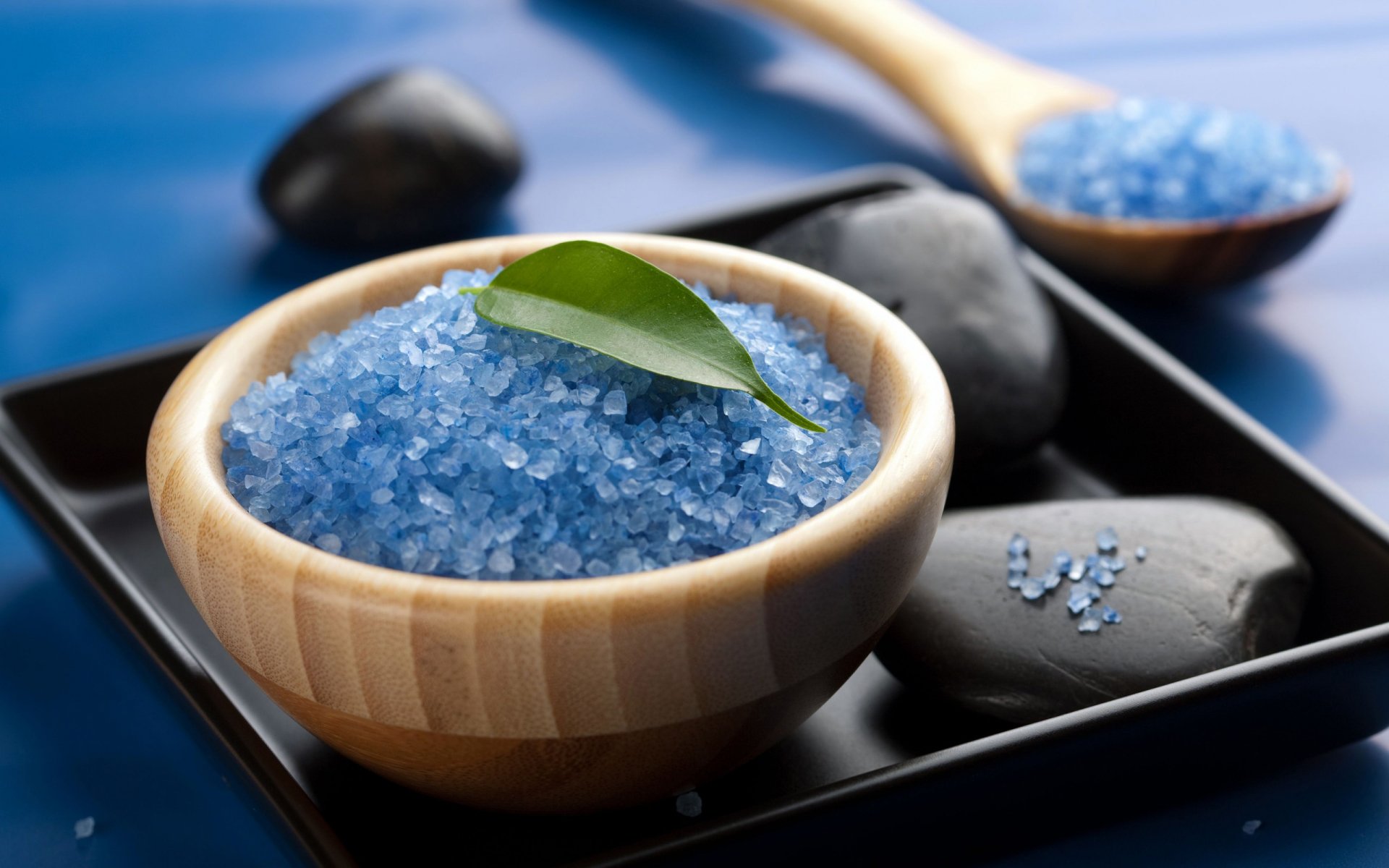 HD desktop wallpaper of blue spa salts in a wooden bowl with black stones and a green leaf, creating a calming, man-made spa atmosphere.