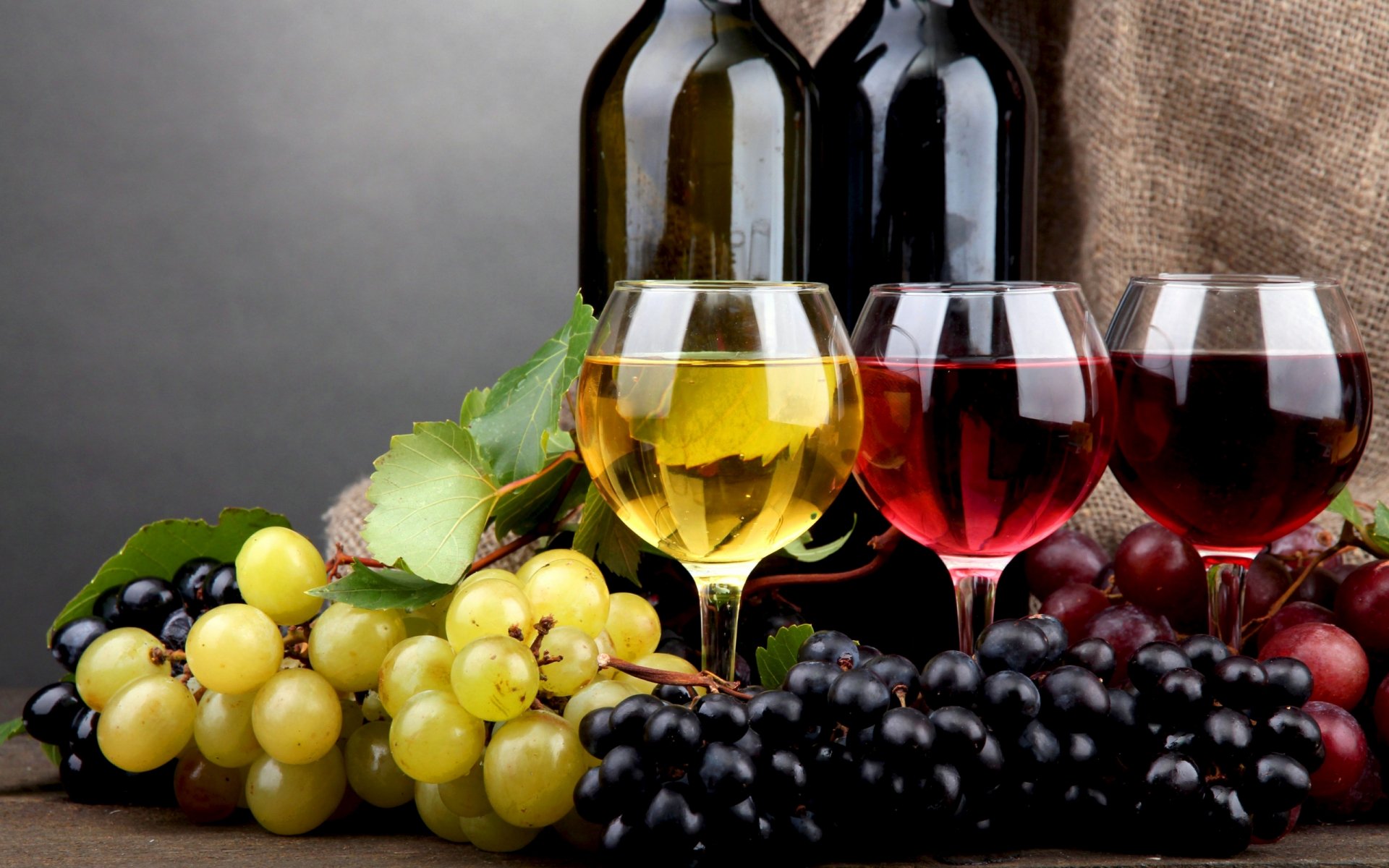 HD PC desktop wallpaper featuring food and wine with glasses of white, rosé, and red wine surrounded by fresh green, red, and black grapes against a neutral background.