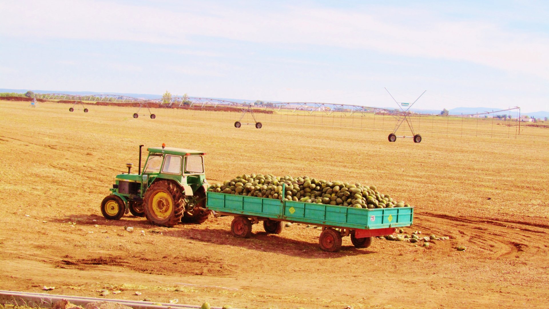4K Ultra HD desktop wallpaper featuring a green tractor pulling a trailer loaded with produce across a vast, arid farmland under a clear sky.