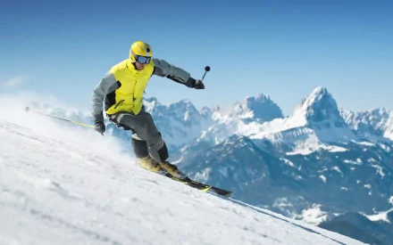 Action shot of a skier in a bright yellow jacket carving a snowy slope with alpine peaks in the background — 2K Quad HD PC desktop wallpaper (sports, skiing)