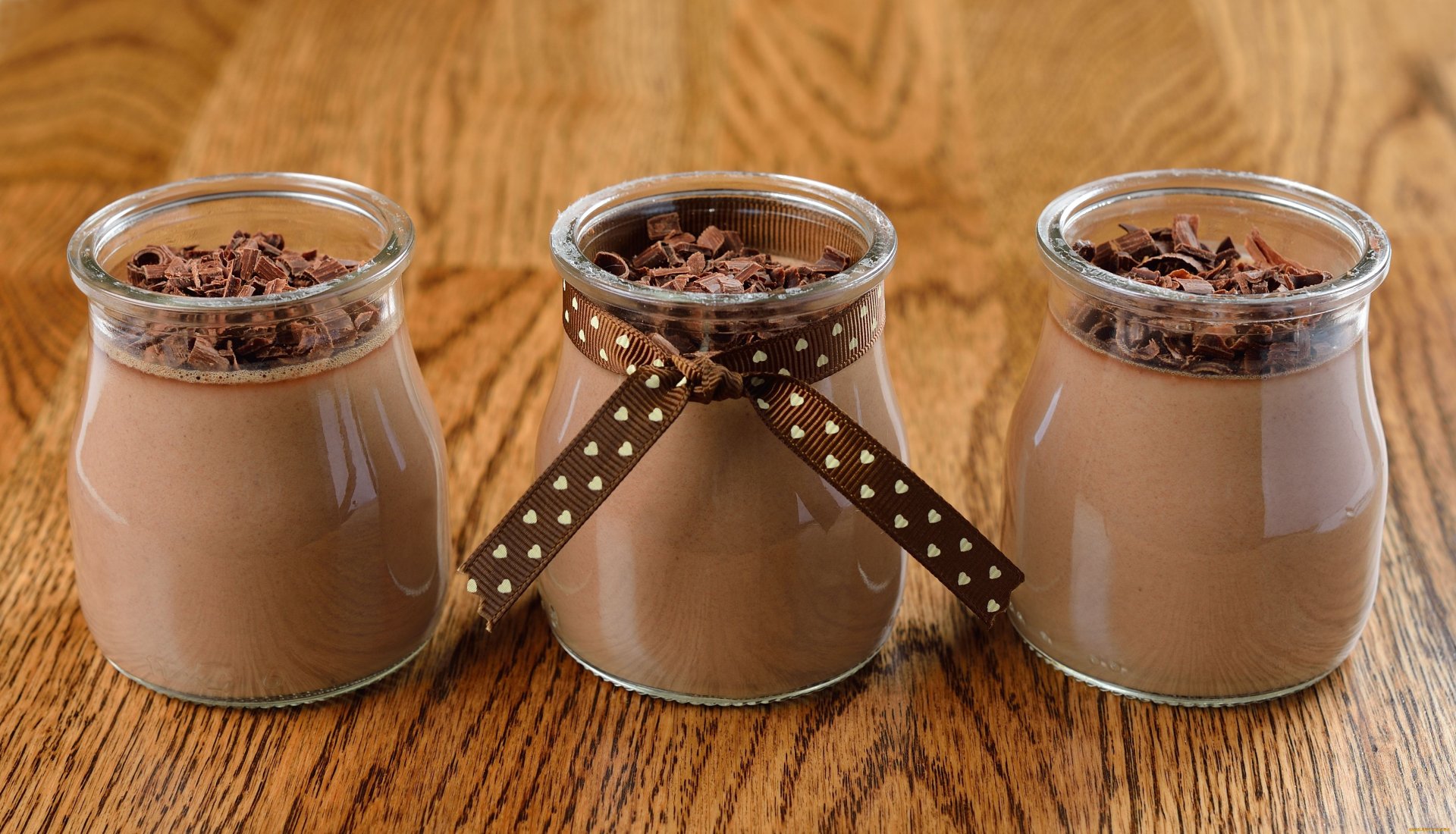 Three jars of creamy chocolate dessert topped with chocolate shavings, set against a warm wooden background. This 4K Ultra HD image captures a delightful food moment.