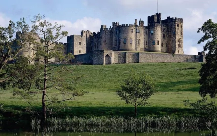 HD PC desktop wallpaper showing a man-made Northumberland castle on a grassy hill beside a reflective pond, framed by trees under a blue sky.