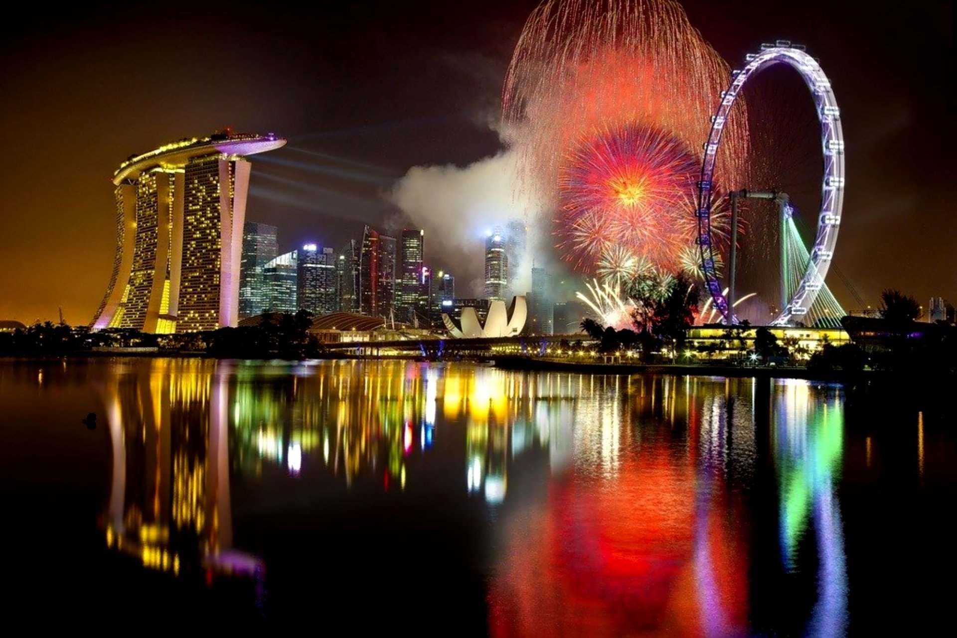 HD PC desktop wallpaper background — night photography of a city skyline with vibrant fireworks bursting over the waterfront, colors and lights reflected in the calm water.