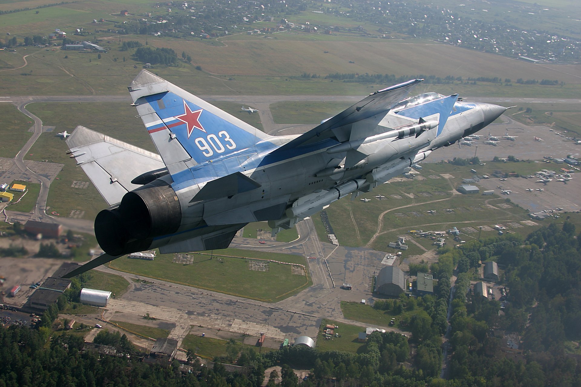 HD desktop wallpaper featuring a military Mikoyan MiG-31 jet soaring above a landscape with visible buildings and open fields below.