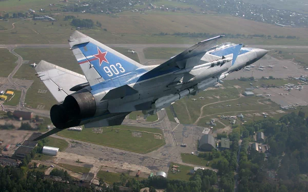 HD desktop wallpaper featuring a military Mikoyan MiG-31 jet soaring above a landscape with visible buildings and open fields below.