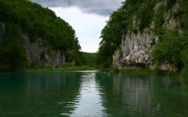 4K Ultra HD PC desktop wallpaper/background: serene river threading a narrow canyon of forested limestone cliffs, emerald water reflecting lush nature beneath a moody cloudy sky.