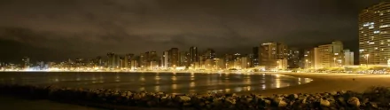 HD desktop wallpaper featuring a city skyline at night, illuminated by golden streetlights and reflected in the calm water, with rocky shoreline in the foreground.