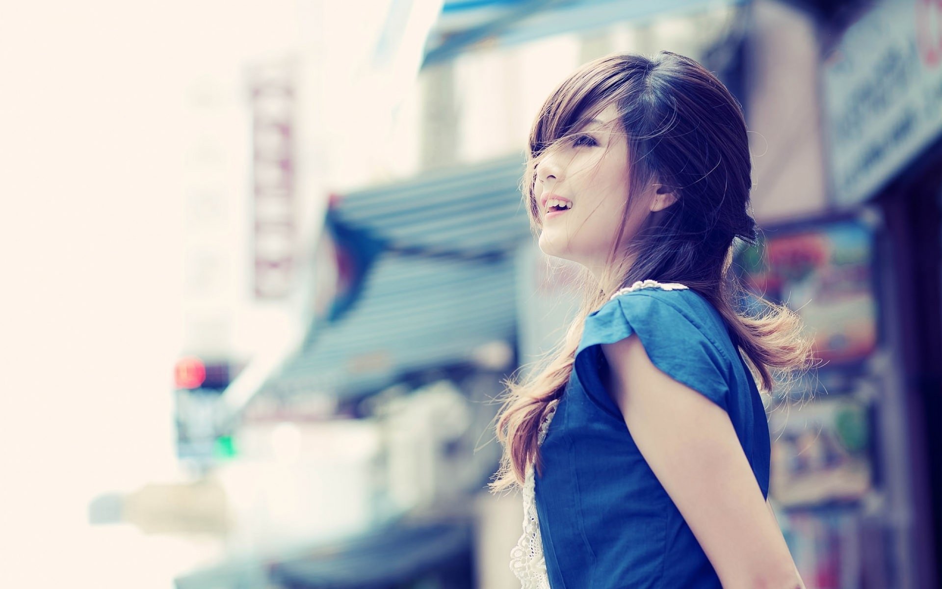 HD desktop wallpaper of an Asian woman smiling, wearing a blue top, standing outdoors with blurred city elements in the background.